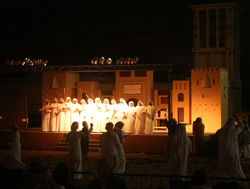 Emirati Men performing the Yowla, a traditional dance in the heritage of the United Arab Emirates next to a historic building.