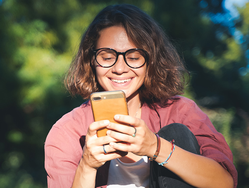Photo of a young woman wearing glasses facing us and smiling as she looks down into her phone.